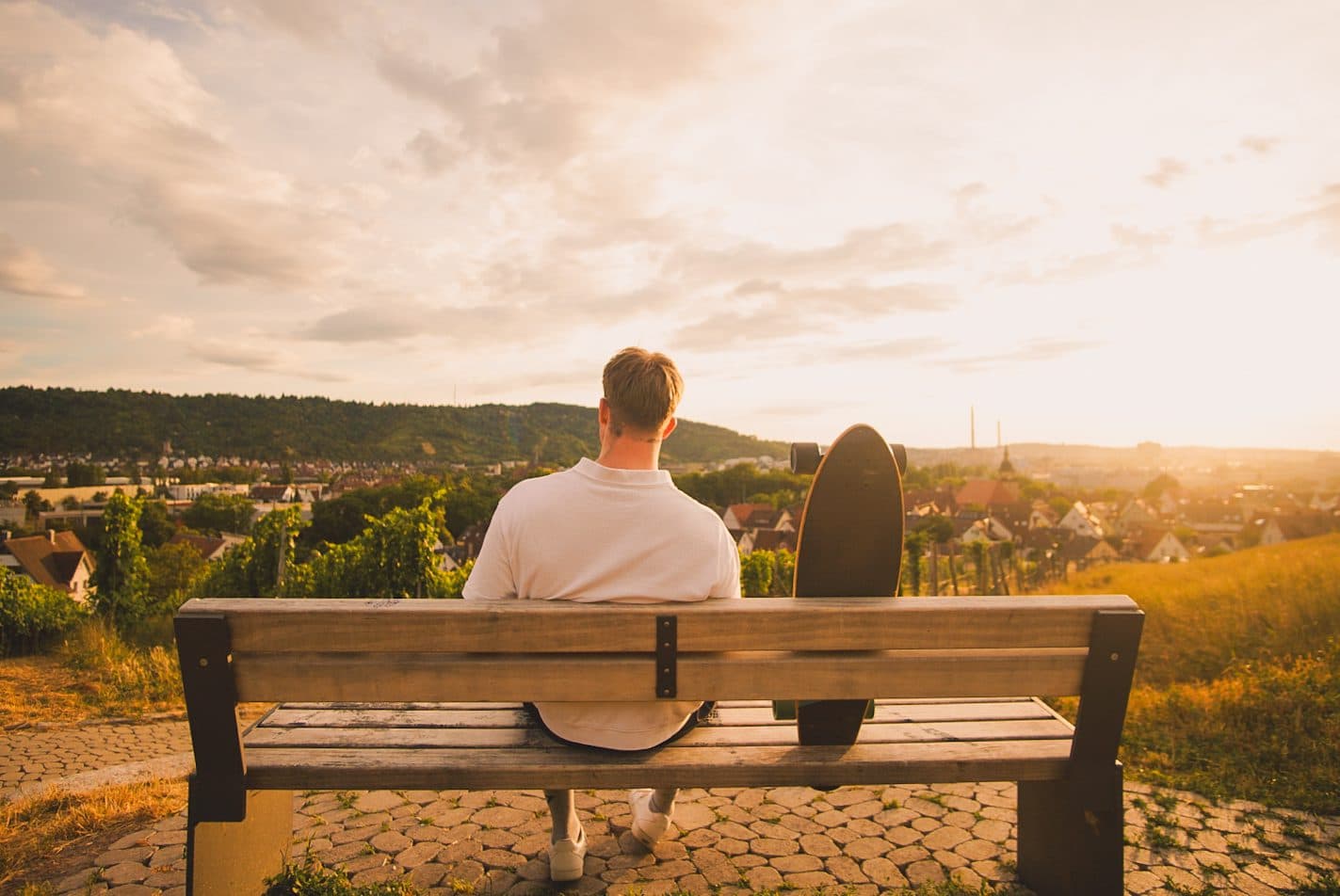 Candy Jack Hoffmann sitzt von hinten auf einer Bank über den Weinbergen, daneben ein Longboard, Sonnenuntergang über Stuttgart